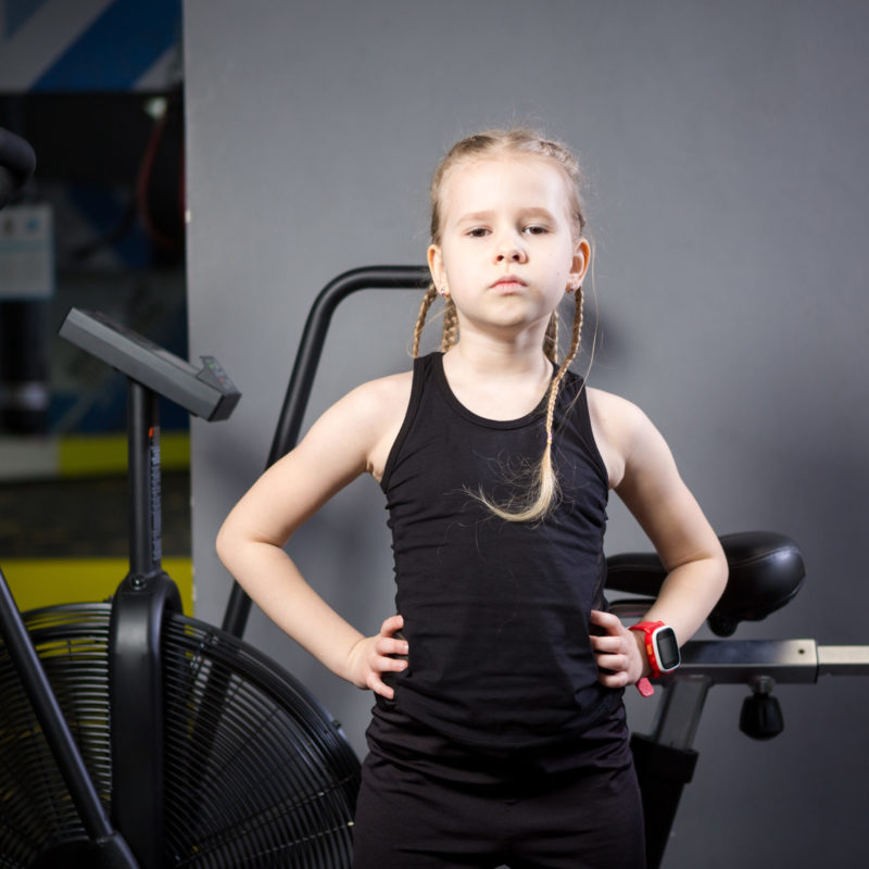 Small attractive caucasian child using exercise bike in the gym. Fitness. A little athlete using an air bike for a cardio workout at the crossfit gym. Athletic girl posing near the cycling machine.